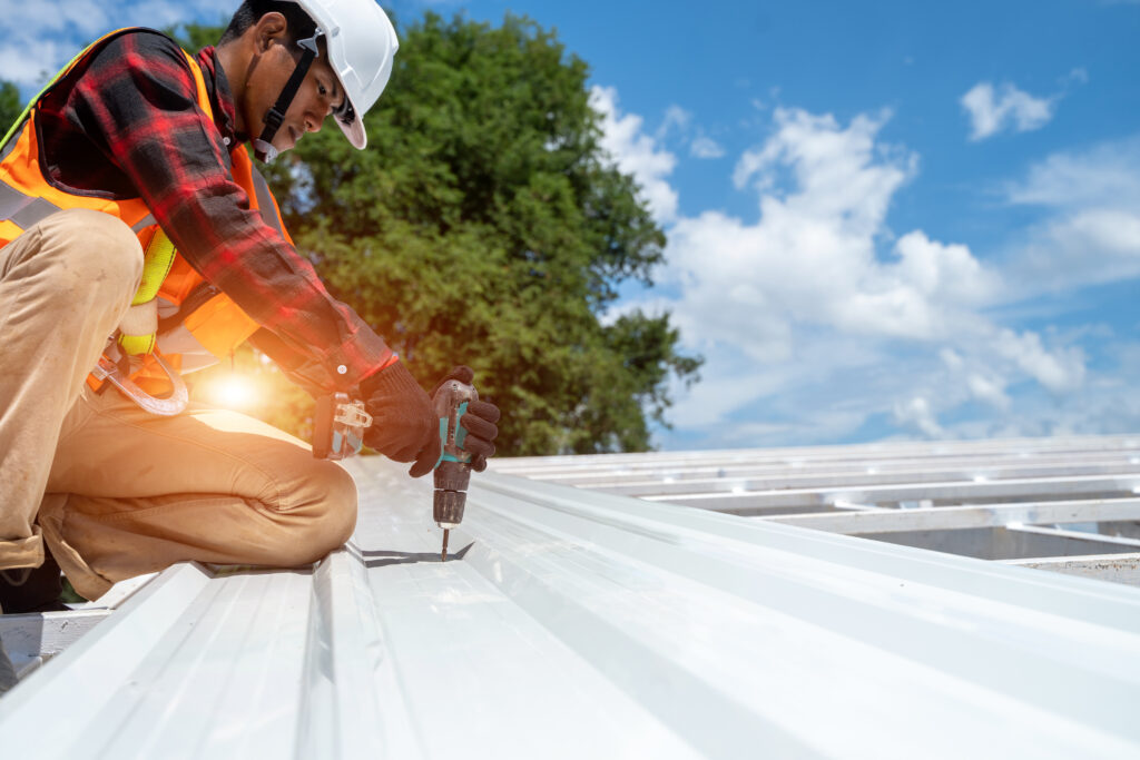 A professional roofing contractor applies elastomeric roof coating to a metal roof, using a drill under a sunny sky with scattered clouds, emphasizing durability and leak prevention.